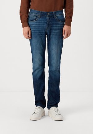 Blauwe denim jeans met een rechte pijp, vijf zakken en subtiele vervaging. Gecombineerd met witte sneakers en een bruine blouse met lange mouwen.