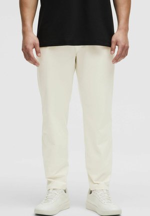 Svetlo bež elastični hlače s tapered krojem, izdelane iz gladkega materiala in s stranskimi žepi, v kombinaciji z belimi supergami.