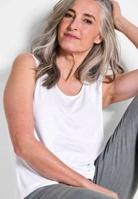Weißes ärmelloses Oberteil mit rundem Ausschnitt, aus einem glatten, leichten Stoff gefertigt. Das Model hat lange, leicht gewellte graue Haare. Graue Hose sichtbar.