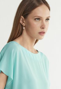 Ljus tealblus i en luftig, lös design med rund halsringning. Har mjukt tyg och korta ärmar. Pärldroppörhängen som accent.