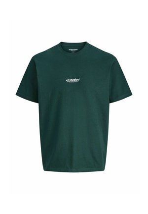 Ung person med kort hår iført en mørkegrøn JJ Studios t-shirt, løse blå jeans og brune sko står mod en ensfarvet lys baggrund.