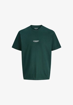 Jonge persoon met kort haar, die een donkergroen JJ Studios T-shirt draagt, losse blauwe spijkerbroek en bruine schoenen, staand tegen een effen lichte achtergrond.