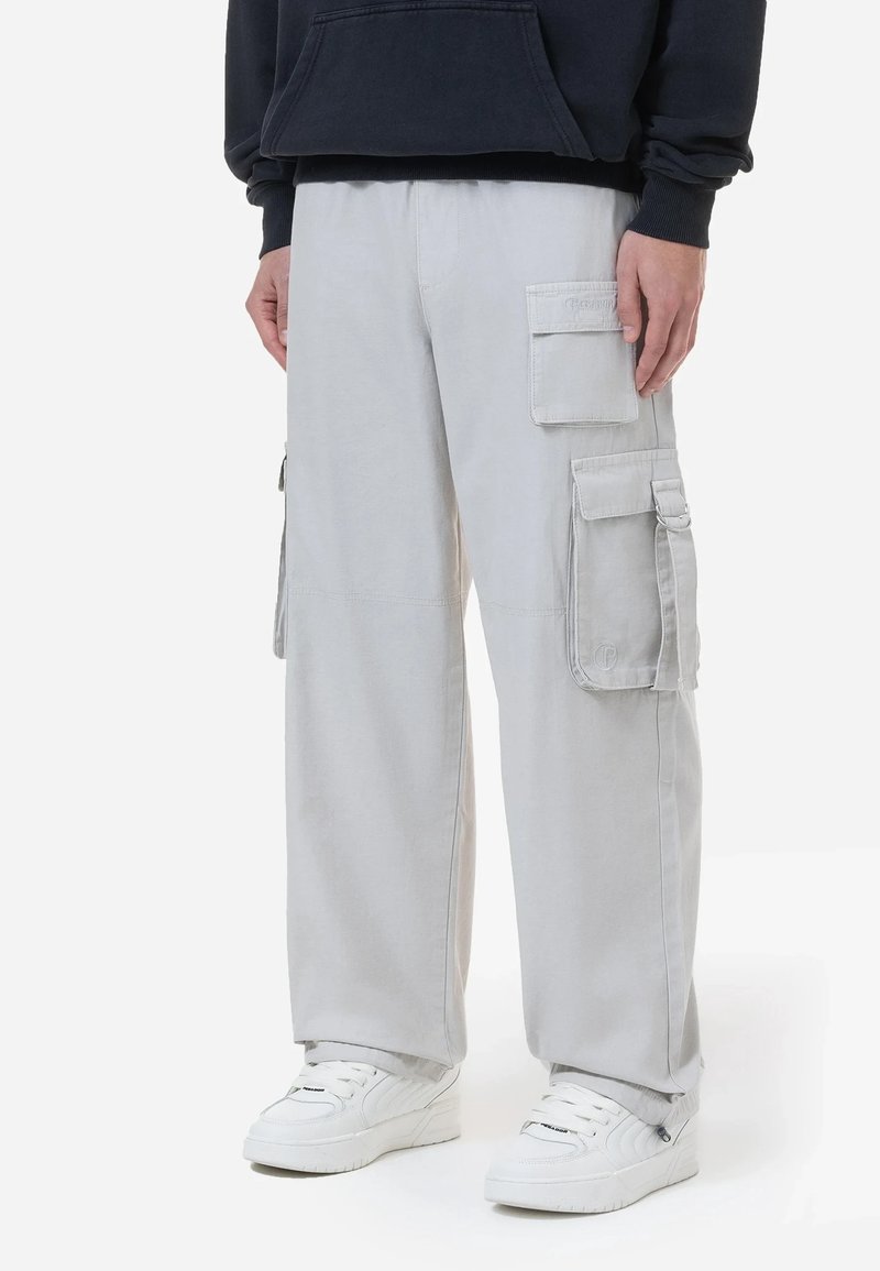 Personne portant un pantalon cargo gris clair avec plusieurs poches, des baskets blanches et un sweat à capuche foncé, debout sur un fond blanc.