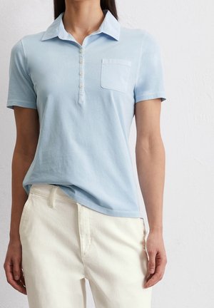 Vrouw draagt een lichtblauw poloshirt met korte mouwen, knopen en een borstzakje, gecombineerd met gebroken witte broek.