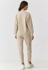 Beżowa bluza z raglanowymi rękawami, ribbowym dekoltem oraz dopasowanymi beżowymi spodniami typu slim-fit. Materiał wydaje się miękki o gładkiej teksturze.