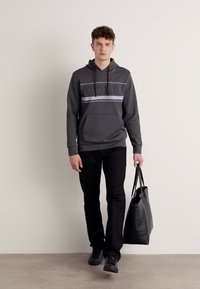 Felpa grigia con logo BOSS testurizzato e striscia orizzontale, abbinata a pantaloni neri e borsa tote nera. Il modello è in piedi su uno sfondo neutro.
