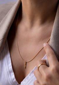 Gouden halsketting met een textuurhanger, voorzien van een slanke ketting en een minimalistisch ontwerp, gedragen over een witte V-hals shirt.