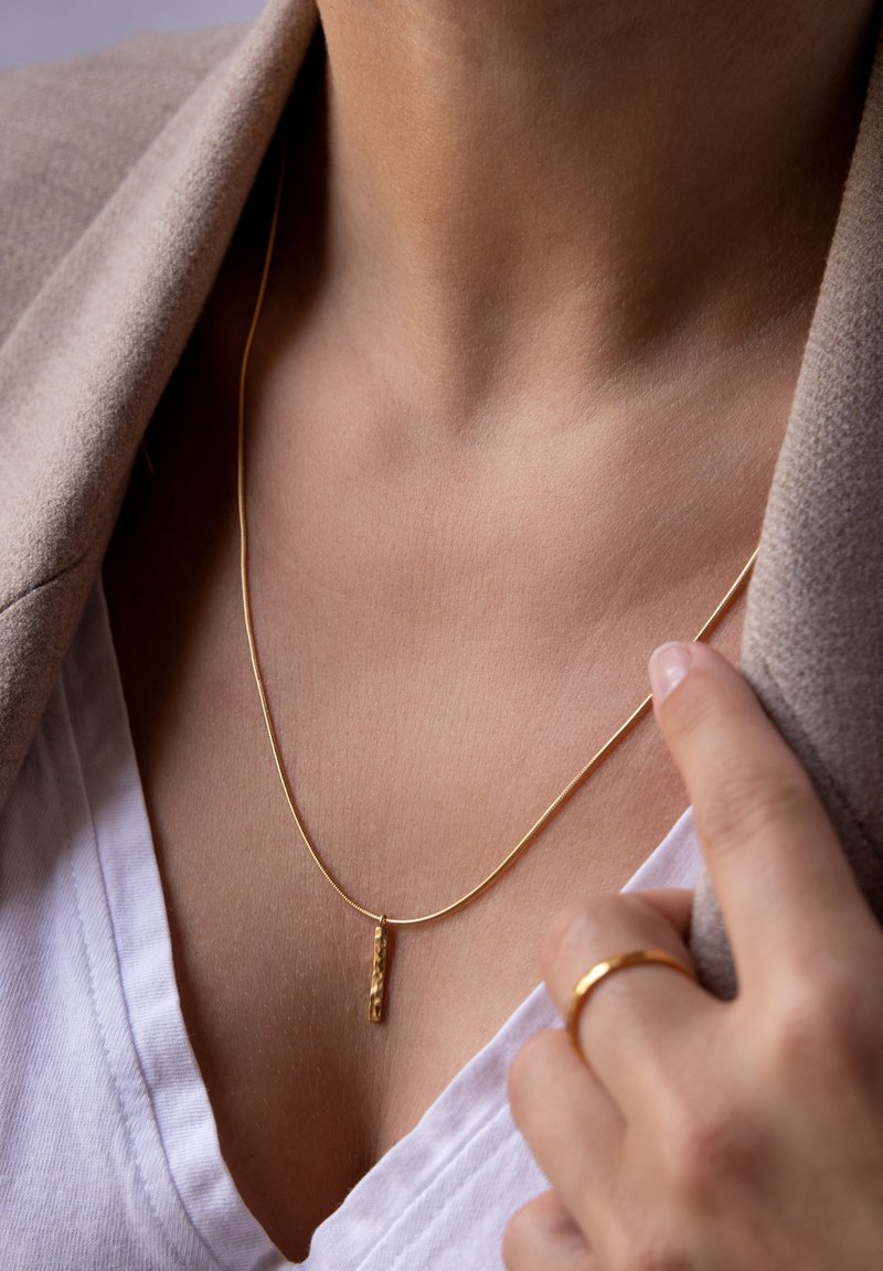 Gouden halsketting met een textuurhanger, voorzien van een slanke ketting en een minimalistisch ontwerp, gedragen over een witte V-hals shirt.