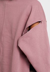 Detailní záběr na rukáv prachově růžové mikiny, který je částečně rozepnutý, přičemž odhaluje ruku uvnitř.