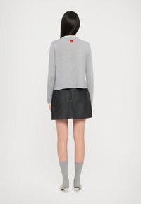 Maglione grigio a maglia con un design cropped, caratterizzato da una toppa rossa sul retro, abbinato a una gonna in denim scuro e calze grigie.