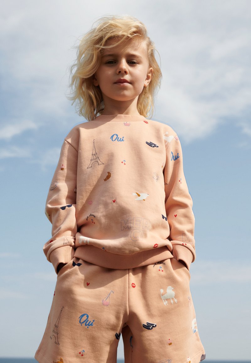 Conjunto de sudadera y pantalones cortos de color durazno que presenta diversos estampados divertidos, incluyendo la Torre Eiffel, pájaros y corazones. Tejido suave, corte relajado.