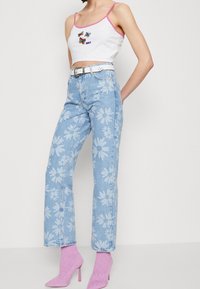 Ingekorte witte tanktop met roze afwerking en vlinderborduursel, gecombineerd met lichtblauwe spijkerbroek met witte bloemenpatronen en roze hakken.