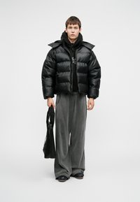 Črna pufasta jakna s sijajnim zaključkom, prevelik kroj, zadrga, oblečena čez črno kapuco. Usklajena s sivimi širokimi hlačami in črno torbo.