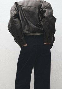 Rjava usnjena jakna z sproščenim krojem, z našitimi detajli, v kombinaciji s temno modrimi kordnimi hlačami, roke v žepih.