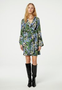 Floral-Wickeldress mit blau, grün und weißen Mustern, langen Ärmeln, einer taillierten Taille und einem ausgestellten Saum. Getragen mit schwarzen kniehohen Stiefeln.