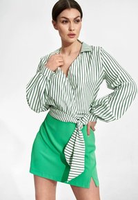 Bluzka z kołnierzykiem w stylu wrap z białymi i zielonymi pionowymi paskami oraz objętościowymi rękawami, w zestawieniu z jasnoniebieską mini spódnicą z bocznym rozcięciem.