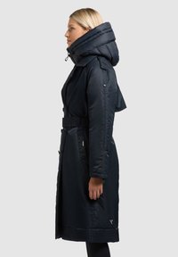 Dolga navtično modra puhasta jakna z velikim kapuco, pasom in stranskimi žepek. Gladek tekstura z nežnim sijajem in logotipom na robu.