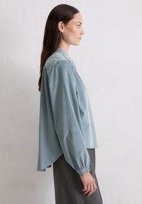 Helles blaues, langärmliges Bluse mit geradem Saum, ausgestattet mit einem hohen Kragen und weichem, strukturiertem Stoff, kombiniert mit maßgeschneiderten grauen Hosen.