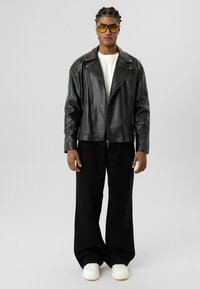 Zwarte leren motorjas over een wit T-shirt, gecombineerd met losse zwarte broek en witte sneakers. De jas heeft asymmetrische ritsen.