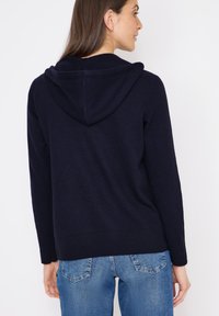 Felpa a maglia blu navy con una texture morbida, maniche lunghe e orlo a costine. Presenta un cappuccio rotondo e una vestibilità casual, abbinata a jeans azzurri.