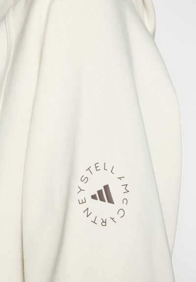 Witte sweatshirtmouw met een rond bruin logo met tekst en drie driehoekige vormen erin. Gladde textuur met een ontspannen pasvorm.
