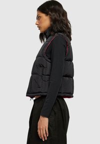 Polstret svart vest med en kort design, med rosa detaljer langs sømmene og høy krage. Lommene på siden og justerbar kant er inkludert.