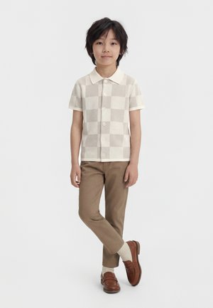 Enfant debout, une jambe croisée, portant une chemise à manches courtes beige à carreaux, un pantalon marron, des chaussettes crème et des mocassins marron.