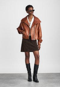 Brun læder bomberjakke med stor krave, hvid snøreshirt, brun suedé mini nederdel og knælange sorte læderstøvler. Sorte solbriller.