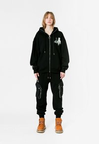 Joven vestido con una sudadera con cremallera negra que tiene un estampado de robot y figura, pantalones de chándal cargo negros con cremalleras, y botas de estilo militar color marrón claro, de pie en posición erguida.