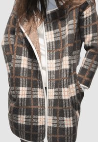 Cardigan de cuadros en gris, marrón y blanco, hecho de un material suave. Presenta un frente abierto, bolsillos laterales y un cuello texturizado.