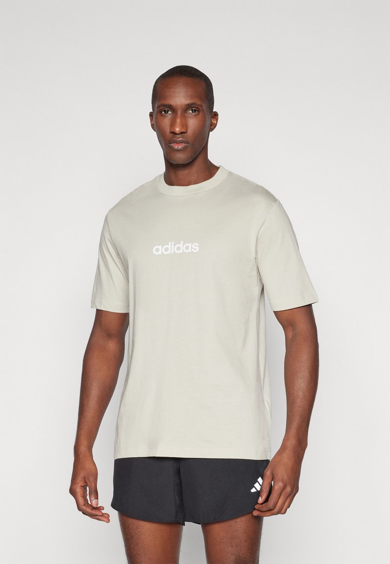 Světle béžové tričko Adidas s kulatým výstřihem a krátkými rukávy, na prsou s bílým logem. Hladká bavlněná látka, jednoduchý design.
