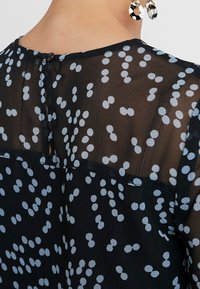Durchsichtige schwarze Bluse mit weißen Punkten und einer kleinen Knopfverschluss hinten oben, getragen mit schwarz-weißen runden Ohrringen.