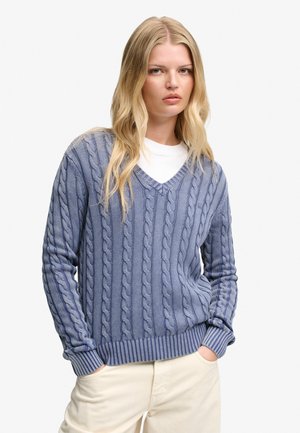 Blonde vrouw met een lichtblauwe kabelgebreide V-hals trui over een wit overhemd en crèmekleurige broek, staand met handen in de zakken.
