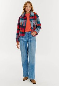 Károvaná bunda v rôznych farbách z vlnenej zmesi, so veľkými gombíkmi a dvoma prednými vreckami. Skombinovaná s oranžovým sveterom a širokými modrými džínsami.