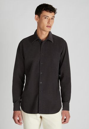 Persona vistiendo una camisa negra de botones de manga larga y pantalones de color claro, de pie contra un fondo gris claro liso.