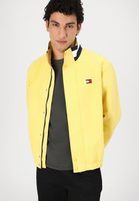 Gelbe Jacke mit Druckknöpfen, hohem Kragen und einem Marineakzent am Halsausschnitt. Besitzt ein kleines Logo-Patch auf der Brust. Getragen über einem grauen Hemd.