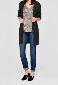 Cardigan lungo grigio scuro sopra una blusa con motivo floreale, abbinato a jeans blu arrotolati e mocassini neri, con una vestibilità comoda e tasche.