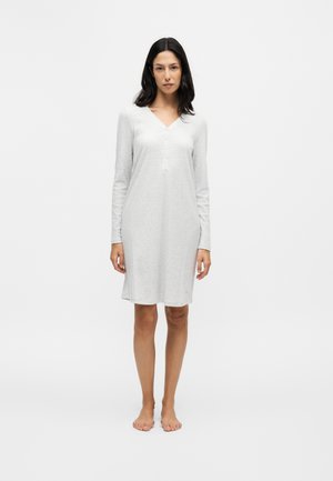 Vrouw met lang donker haar, gekleed in een lichtgrijze jurk met lange mouwen tot op de knie, staande op blote voeten tegen een effen witte achtergrond.