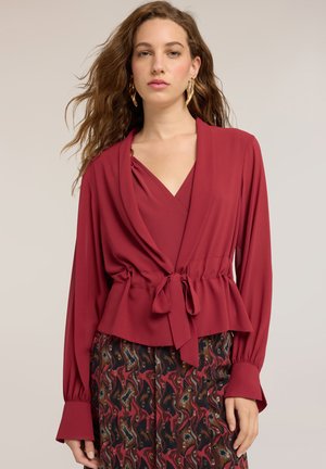 Kvinde med bølget brunt hår iført en dyb rød bluse med lange ærmer og bindebånd foran samt et mønstret skørt med et abstrakt design i rødt, sort og beige.
