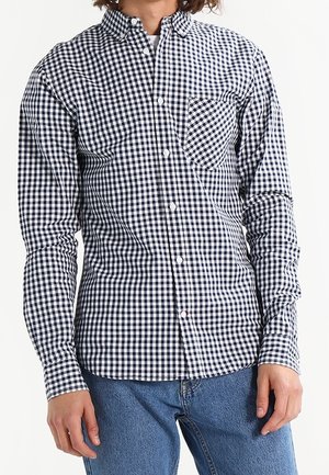 Joven con camisa de cuadros en blanco y negro de manga larga con botones y vaqueros azules sobre un fondo liso.