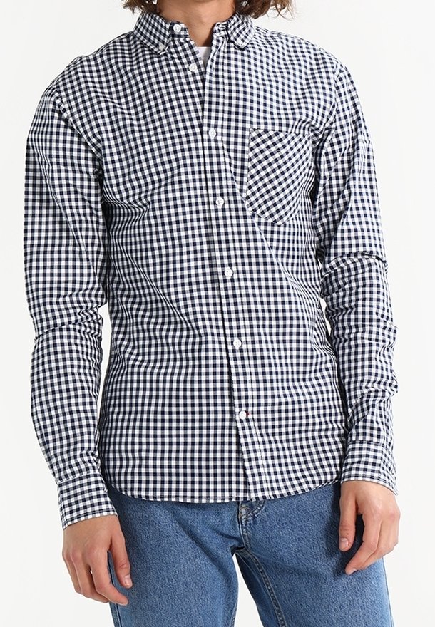 Jeune personne portant une chemise à manches longues à carreaux noirs et blancs boutonnée et un jean bleu, sur un fond uni.