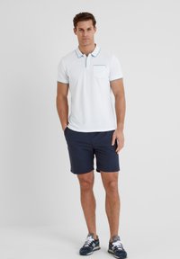 White polo shirt with turquoise accents and a front pocket, paired with navy shorts. Model stands with arms relaxed at sides.