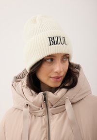 Kremowa prążkowana dzianinowa czapka z kontrastującym czarnym haftowanym napisem "BIZUL." Noszona z jasnobeżową puchową kurtką z wysokim kołnierzem.