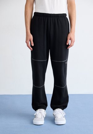 Person iført sorte joggingbukser med elastik i talje og bukseender, hvide sneakers og en hvid skjorte, der står på en blå gulv mod en ensfarvet baggrund.