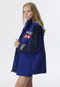 Blaue Jacke aus einer Mischung aus helleren und dunkleren blauen Stoffen, mit mehreren Patches, einem Reißverschluss und Taschen an der Vorderseite.