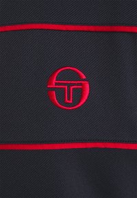 Mörk marinblått tyg med ett strukturerat mönster, med en framträdande röd broderad logotyp och horisontella röda randiga detaljer.