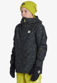 Svart jacka med grön abstrakt mönster, framdragkedja och huva; bärs med en ribbad ljusgul mössa och svarta handskar. Ljusgula byxor.