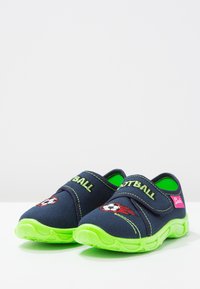 Lichte blauwe stoffen instappers met een groene rubberen zool. Bevat een voetbal afbeelding en groene geborduurde tekst. Klittenbandsluiting.