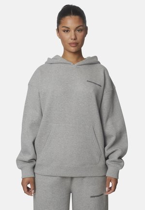 Femme portant un sweat à capuche gris clair et un pantalon de survêtement assorti avec un petit texte noir sur la poitrine et la cuisse.