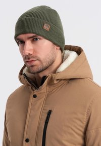 Berretto a coste color verde oliva con una patch in pelle; indossato con una giacca cappuccio tan con colletto foderato in pile e zip nere.